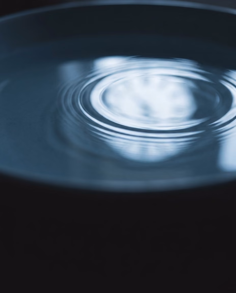 Concentric ripples spreading slowly outward across still deep blue water, a soft circle of window light on the surface