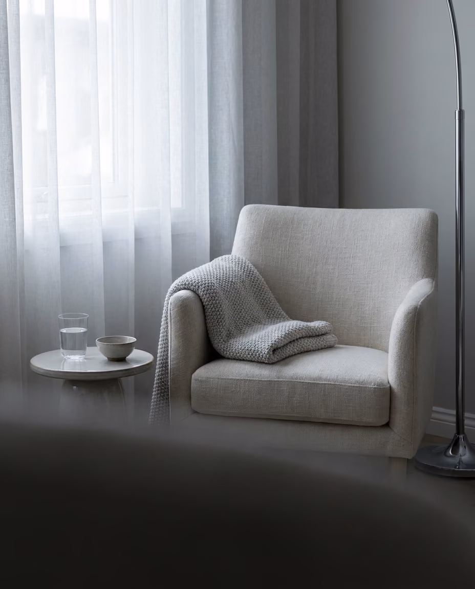 A quiet corner of a home prepared for a treatment: a cream armchair, a folded throw and a glass of water by a curtained window
