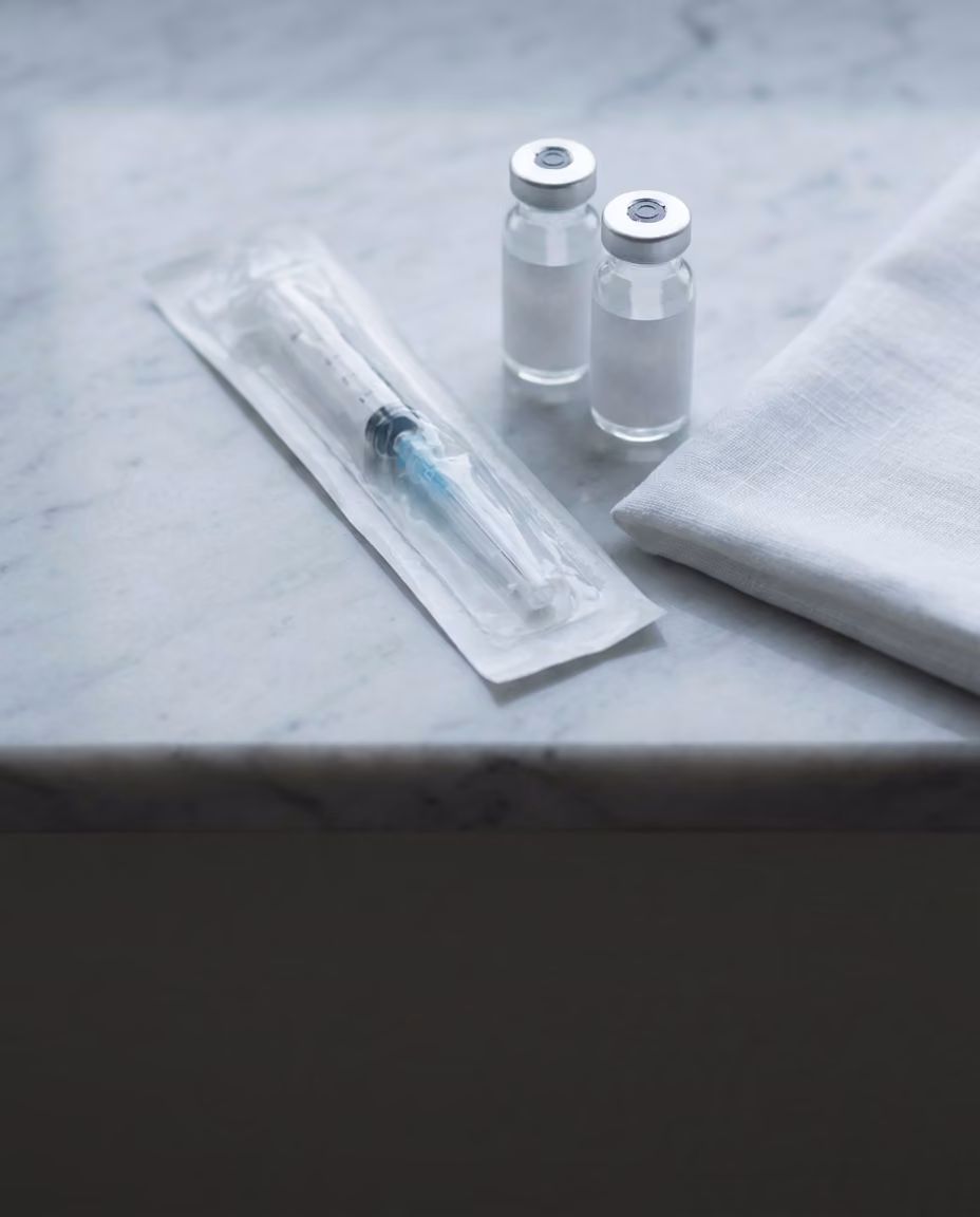 A sealed sterile syringe in its wrapper on pale marble beside two small unlabeled glass vials and a folded white cloth