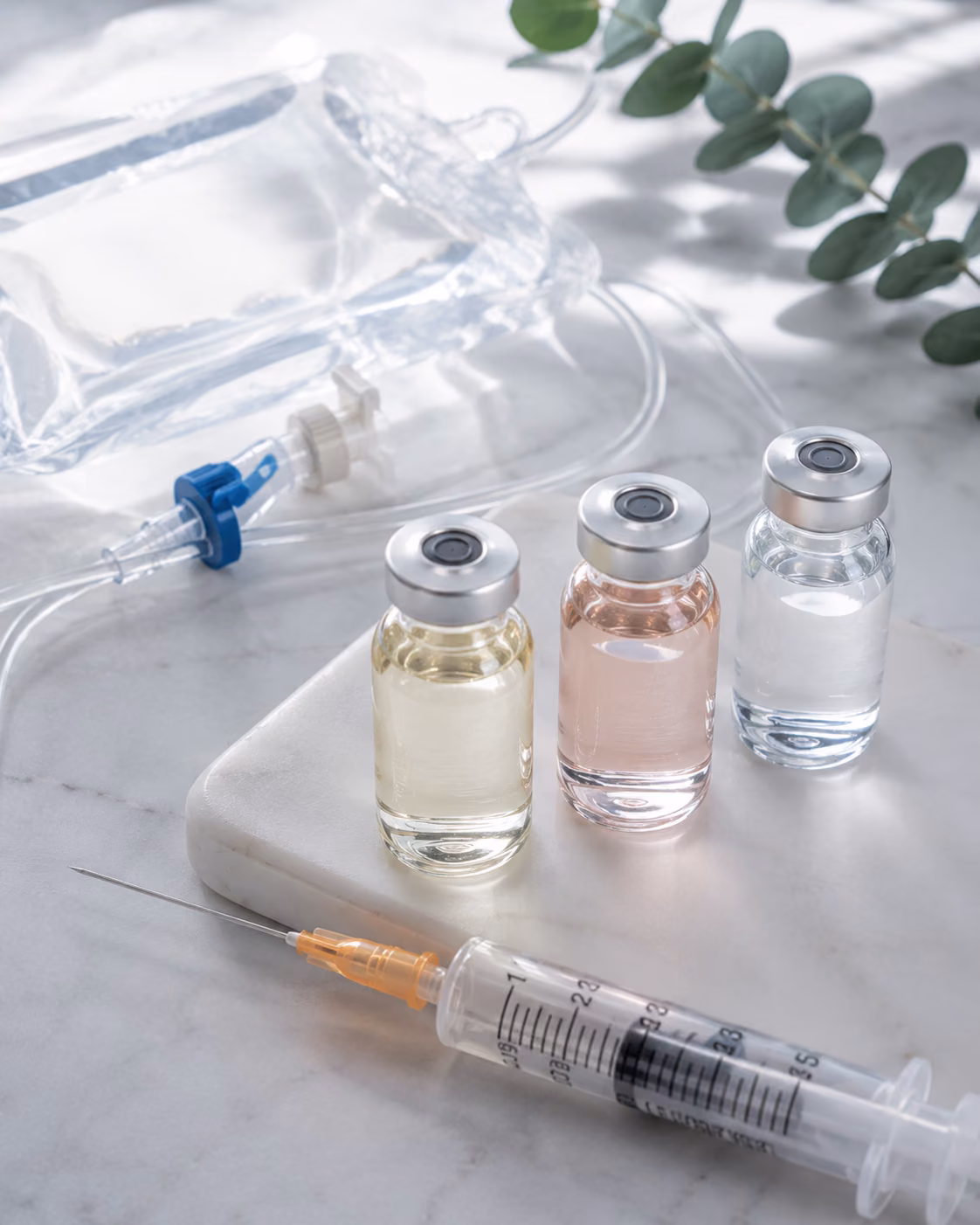 Three unlabeled glass vials of pale yellow, pink and clear fluid on marble, with an intravenous bag, line and a syringe beside them