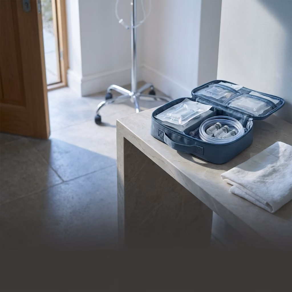 An open medical kit bag set down on a marble console just inside a bright entryway, sealed sterile packs and a coiled line inside, a chrome infusion stand waiting in the room beyond